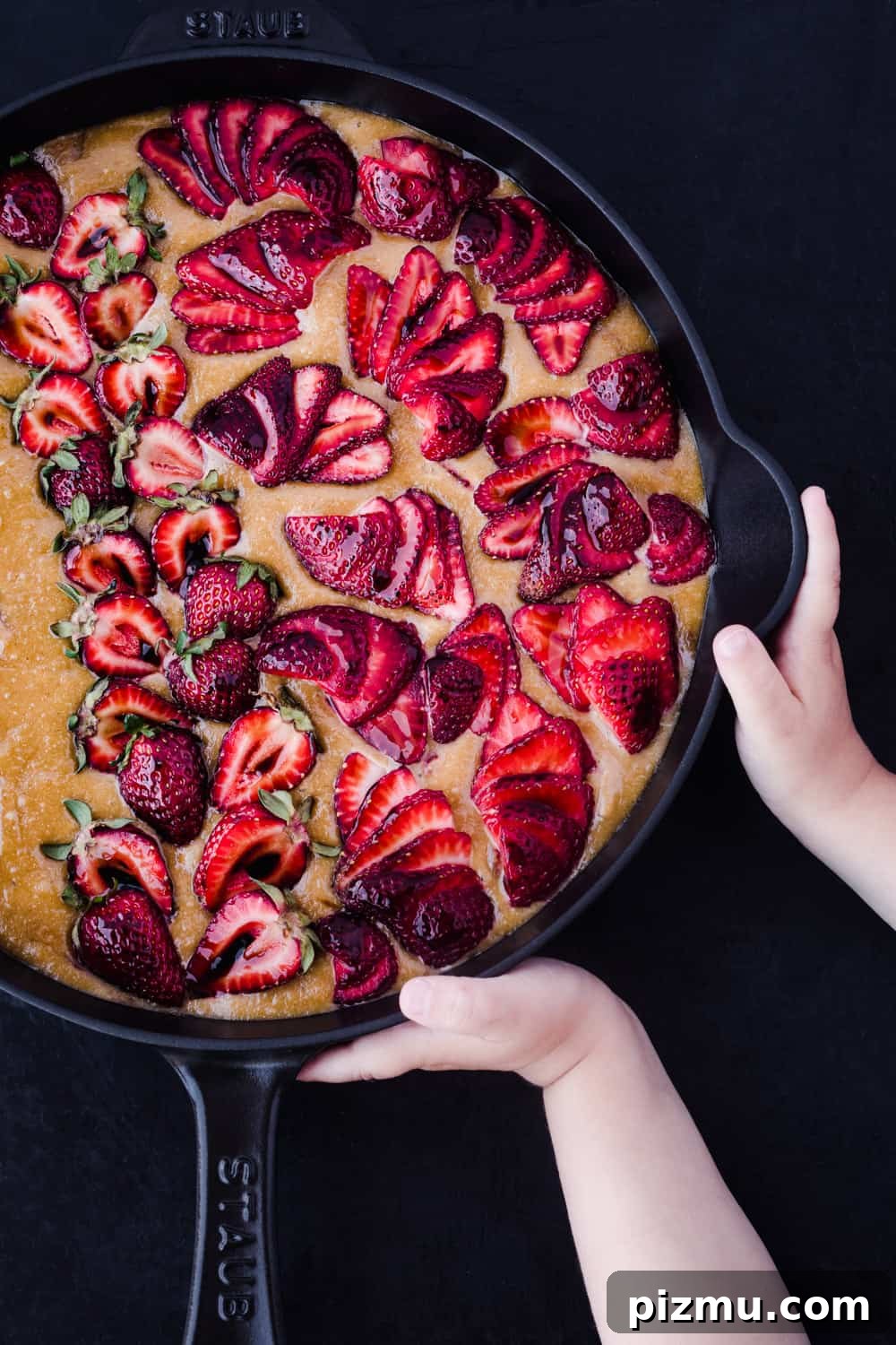 Strawberry Olive Oil Cake for Breakfast, Brunch and Dessert 3 Overhead shot of pre-oven strawberry olive oil cake batter in a Staub cast iron skillet topped with halved fresh strawberries, held by a child's hands on a dark background.