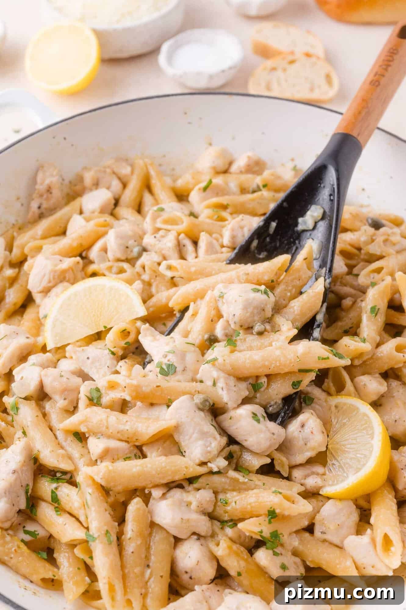 One-Pan Chicken Piccata Pasta with Lemon and Capers 2 A pan of creamy chicken piccata pasta with chunks of chicken, garnished with lemon slices and parsley, being stirred with a black spoon.
