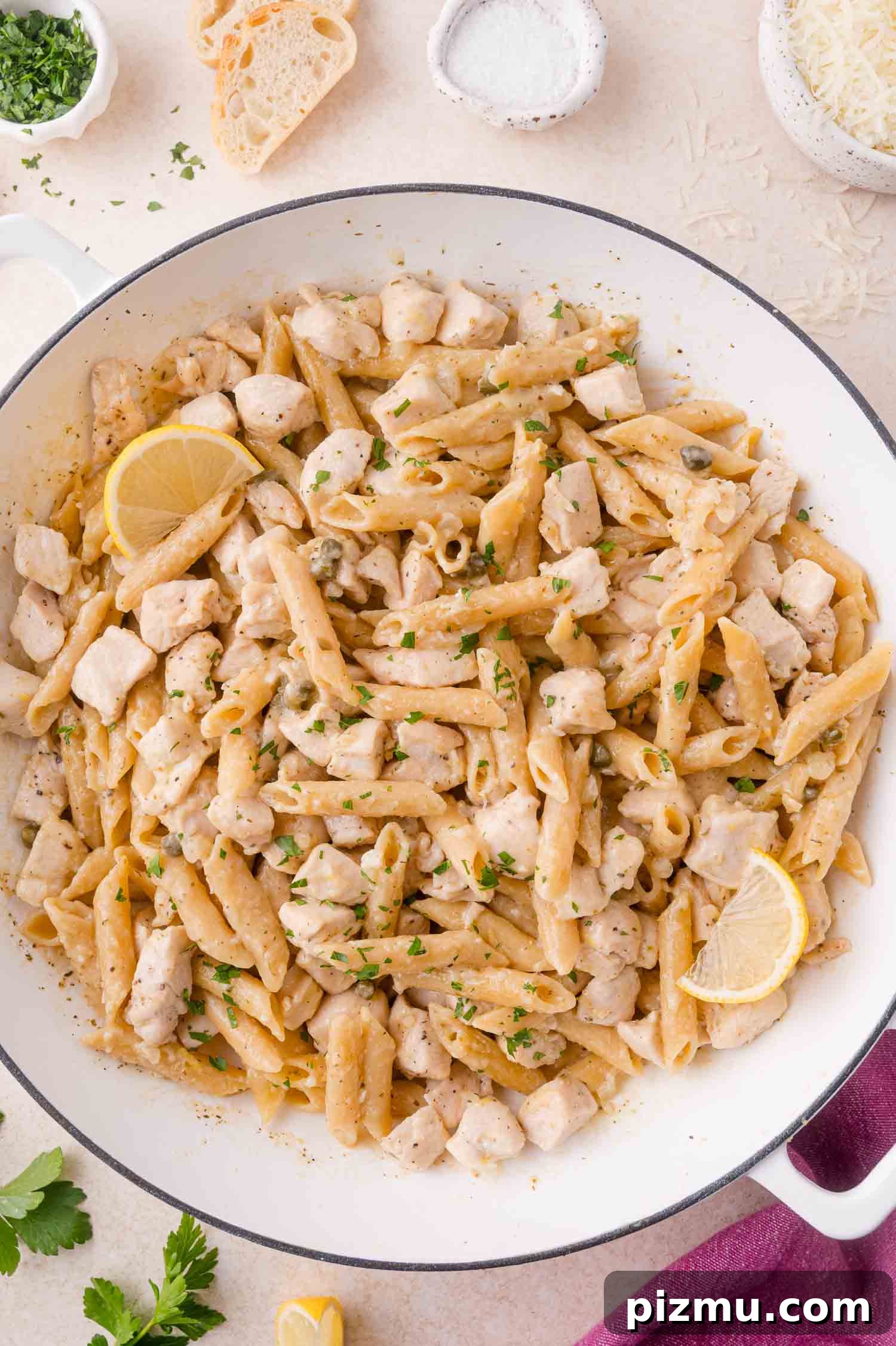 One-Pan Chicken Piccata Pasta with Lemon and Capers 12 A large white pan filled with chicken piccata pasta, with cubed chicken, creamy sauce, chopped herbs, and lemon wedges. Bread slices and seasonings are visible in the background.