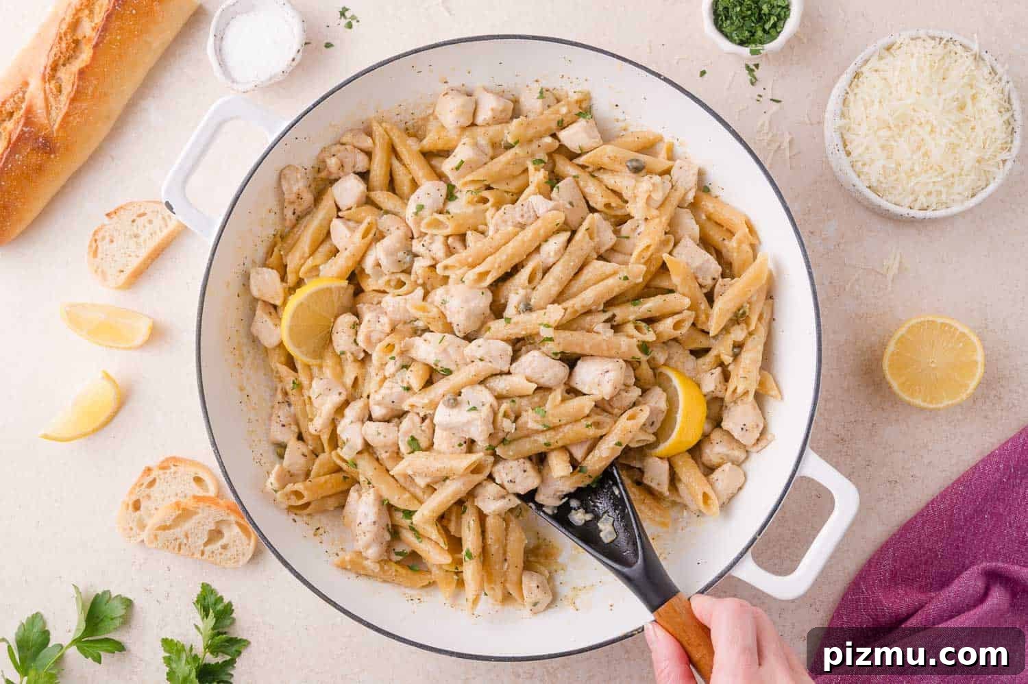 One-Pan Chicken Piccata Pasta with Lemon and Capers 13 A white pan filled with penne pasta, diced chicken, lemon slices, and herbs. A hand holds a black spatula. Bread, cheese, lemon wedges, and parsley are on the table nearby.