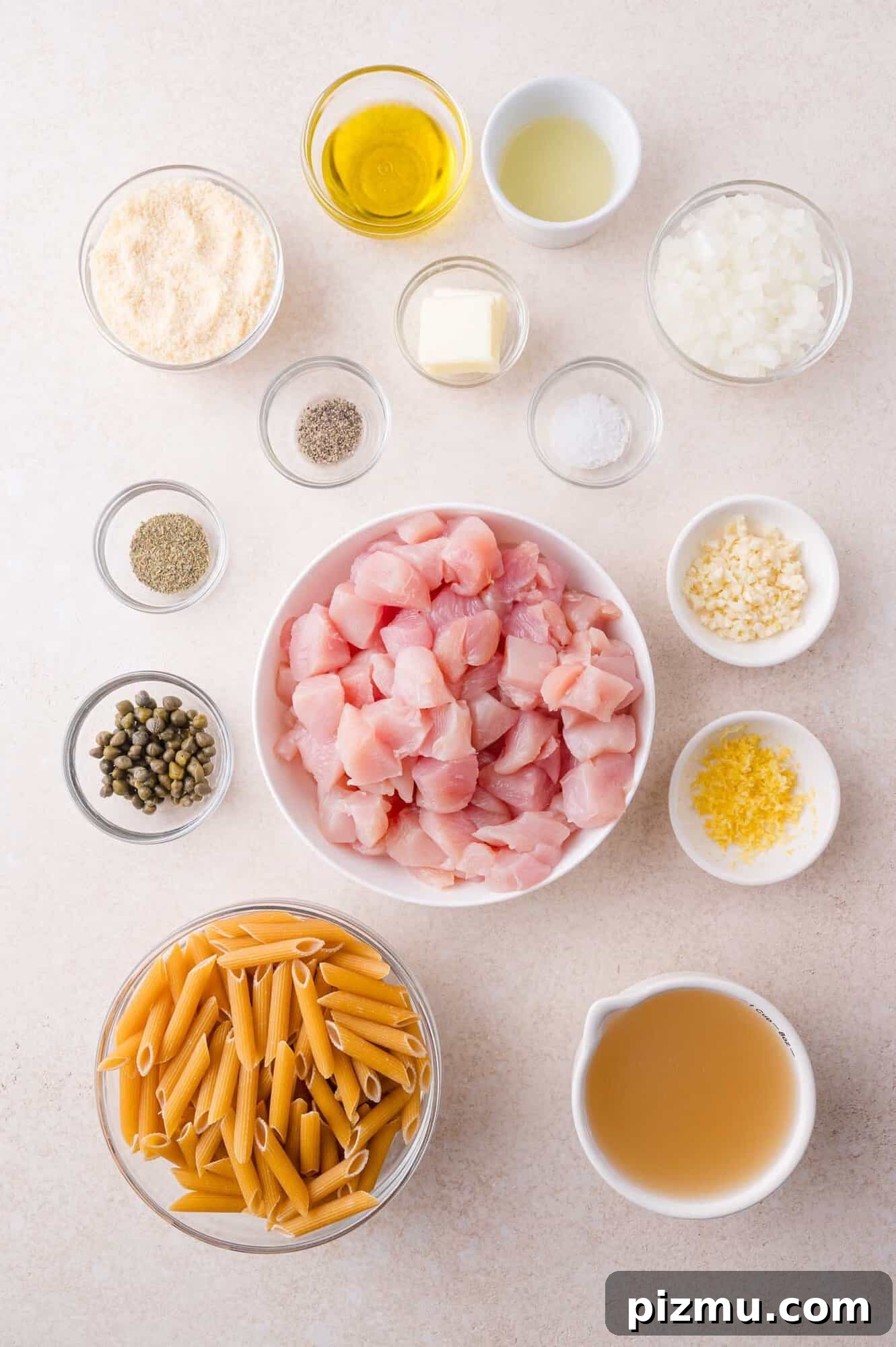 One-Pan Chicken Piccata Pasta with Lemon and Capers 3 Overhead view of ingredients in small bowls on a countertop, including diced chicken, uncooked penne pasta, chopped onion, minced garlic, capers, broth, butter, oil, seasonings, and cheese.