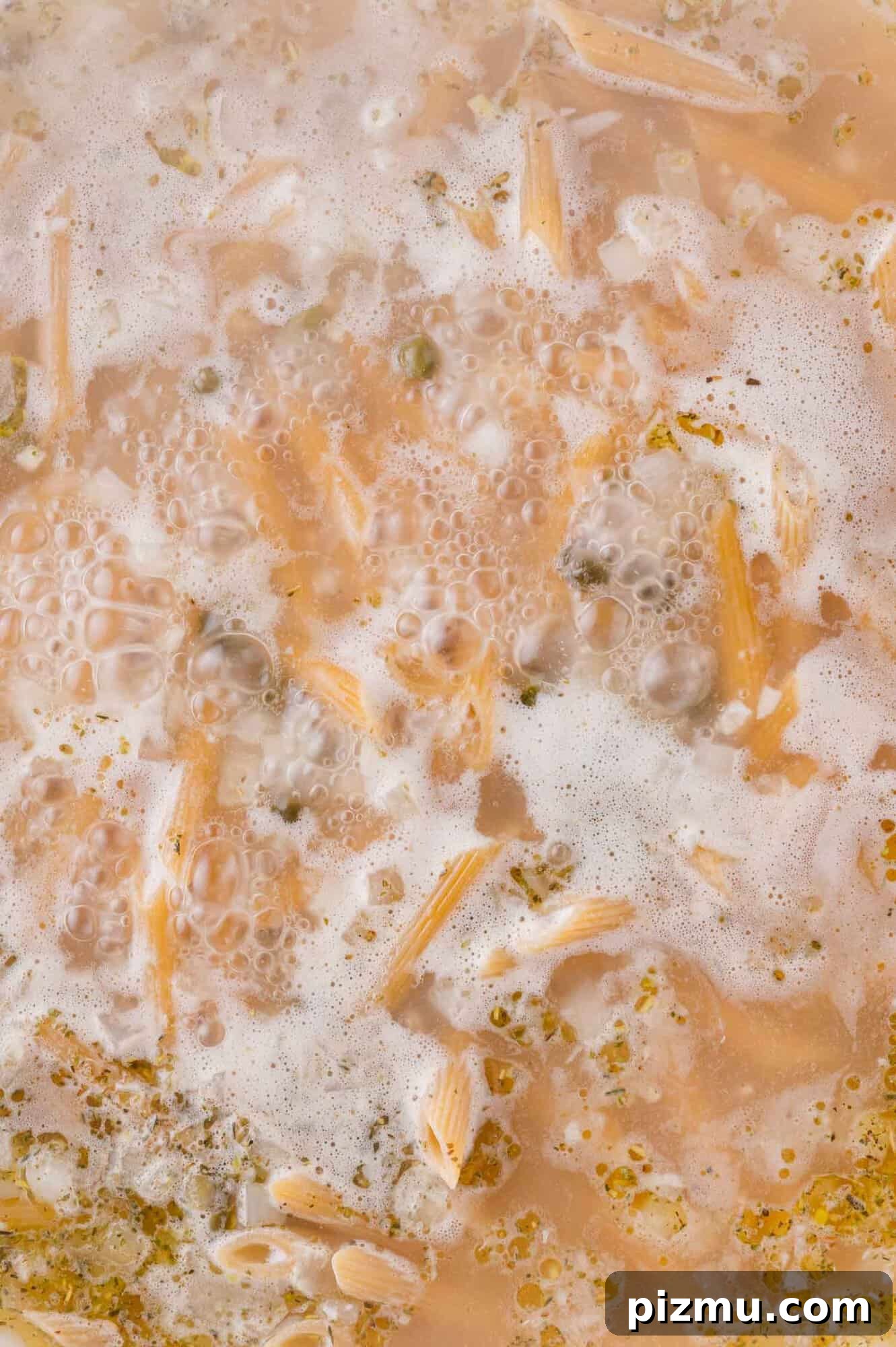 One-Pan Chicken Piccata Pasta with Lemon and Capers 8 Close-up of pasta cooking in boiling water with bubbles, floating foam, and visible herbs and pasta pieces.