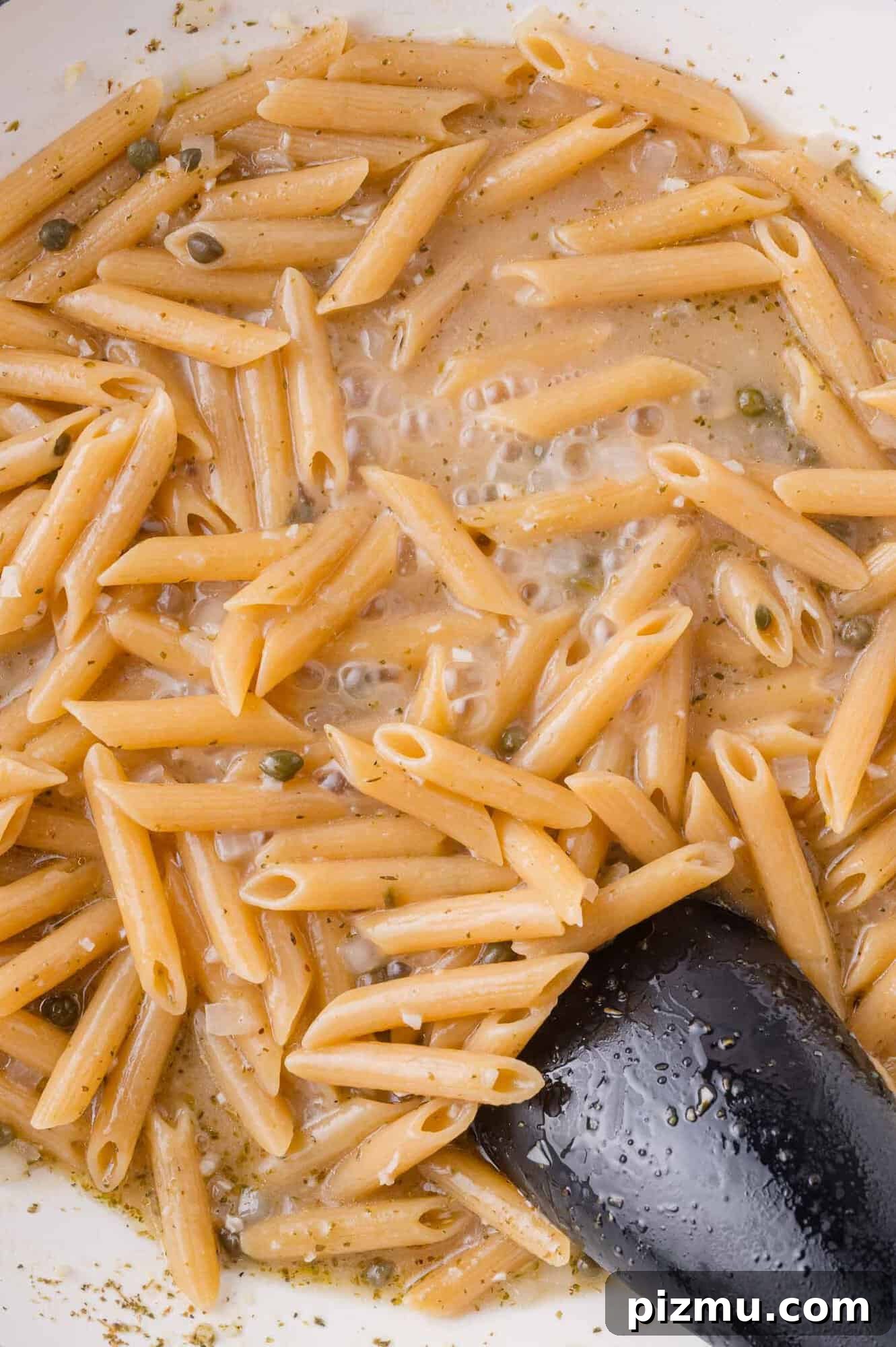 One-Pan Chicken Piccata Pasta with Lemon and Capers 9 Cooked penne pasta in a creamy sauce with visible herbs and capers, stirred with a black serving spoon in a white pan.