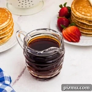 Homemade syrup in a glass pitcher in front of stacks of pancakes with strawberries.