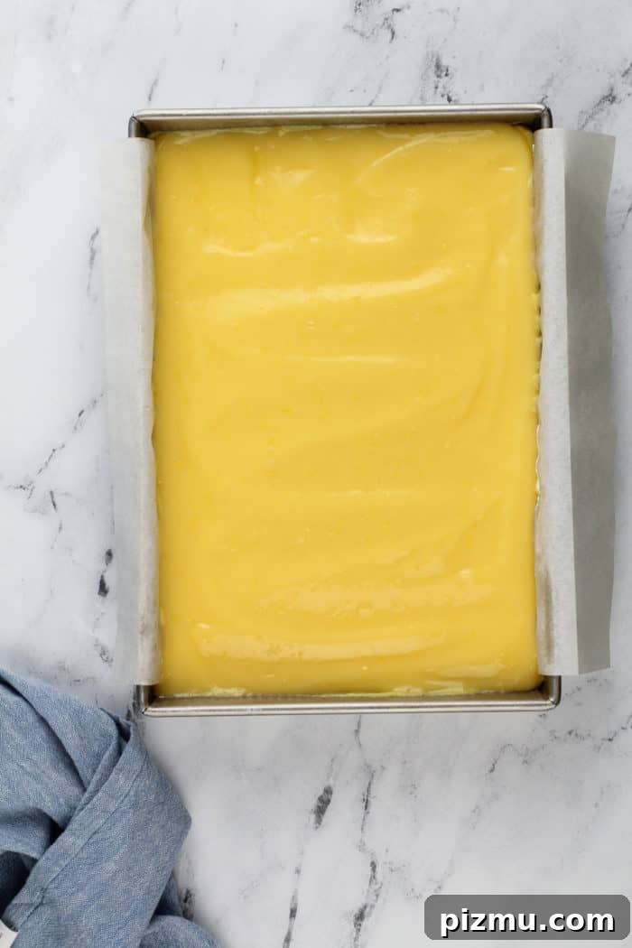 Creamy Layered Lemon Trifle Recipe 9 Lemon curd spread onto layered lemon dessert in a parchment-lined baking pan on a marble countertop.
