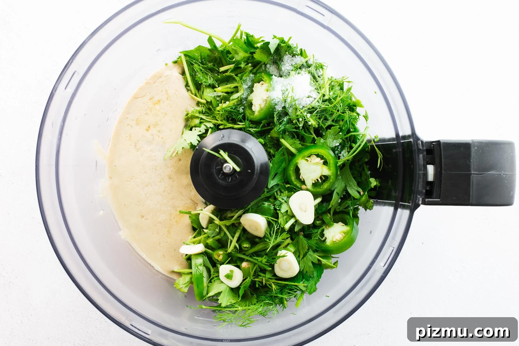 Tahini, fresh herbs, raw garlic and chillies in a food processor as viewed from above.
