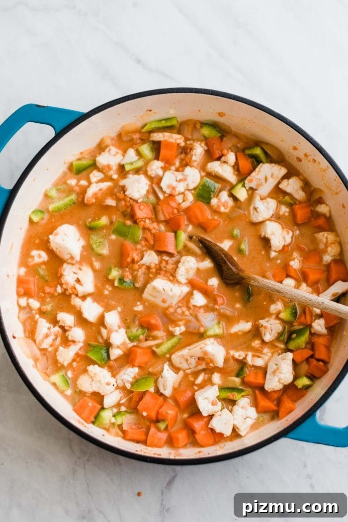 Creamy Red Curry Lentils with Coconut and Spinach 5 Carrot, cauliflower, celery and onion cooking in a red curry broth in a large pot. A wooden spoon rests in the pot.