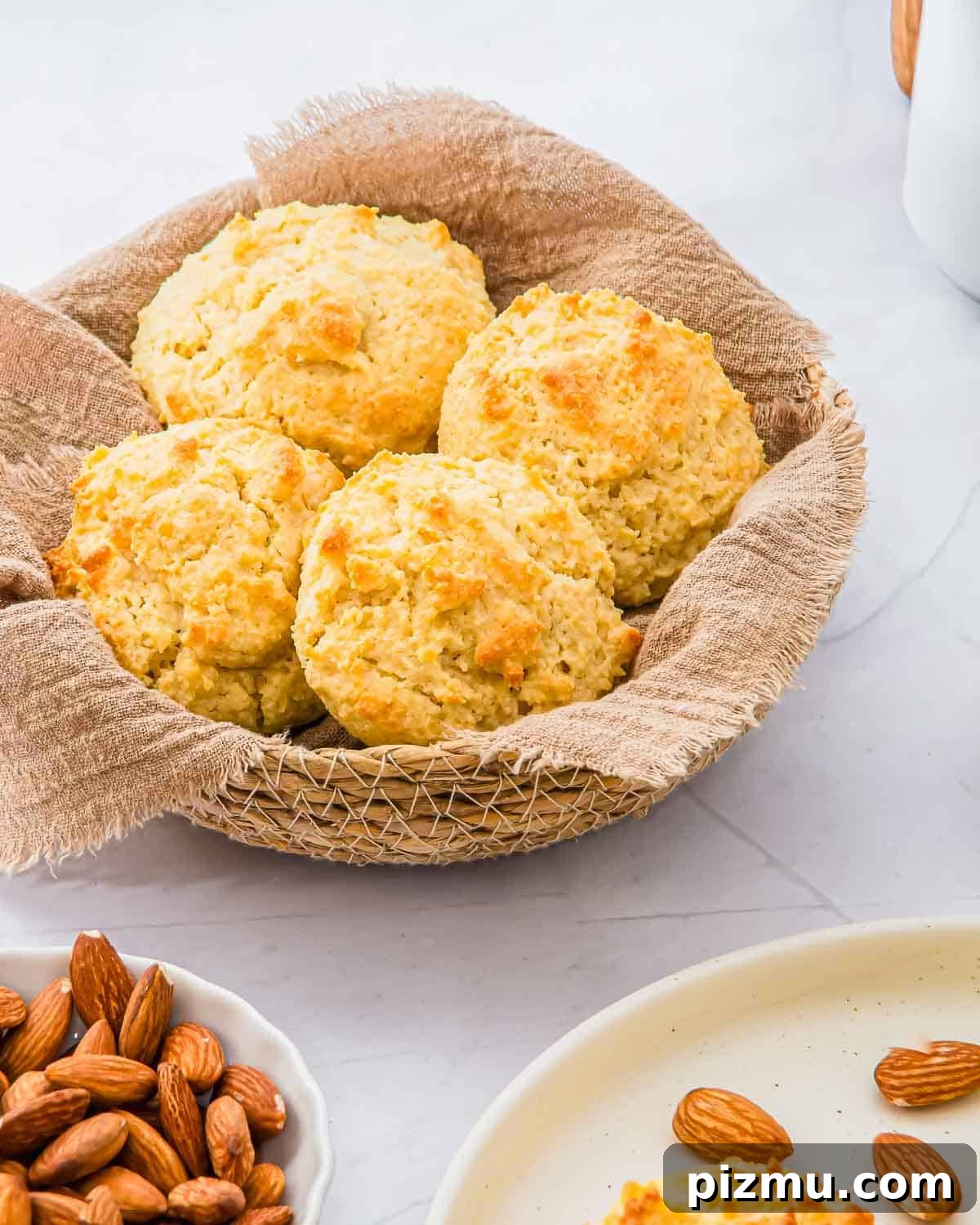 Flaky Almond Flour Biscuits Recipe 2 A basket lined with brown cloth holds four golden, freshly baked almond flour biscuits. Nearby, a small plate of whole almonds and part of a white plate are visible on a light marble surface.