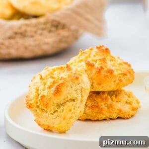 Flaky Almond Flour Biscuits Recipe 12 Two golden-brown drop biscuits sit stacked on a white plate, with a basket of more biscuits in the background. The biscuits have a crisp, textured surface and a light, fluffy appearance.
