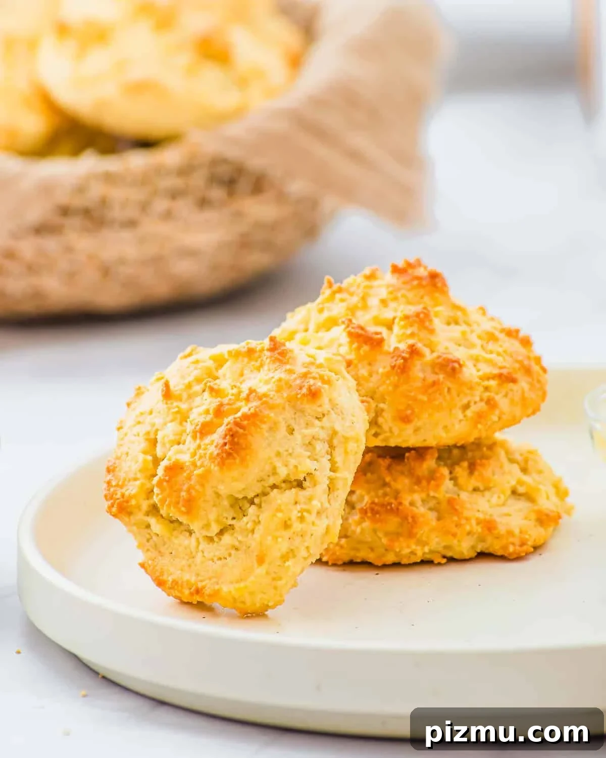 Flaky Almond Flour Biscuits Recipe 3 Two golden-brown drop biscuits sit stacked on a white plate, with a basket of more biscuits in the background. The biscuits have a crisp, textured surface and a light, fluffy appearance.