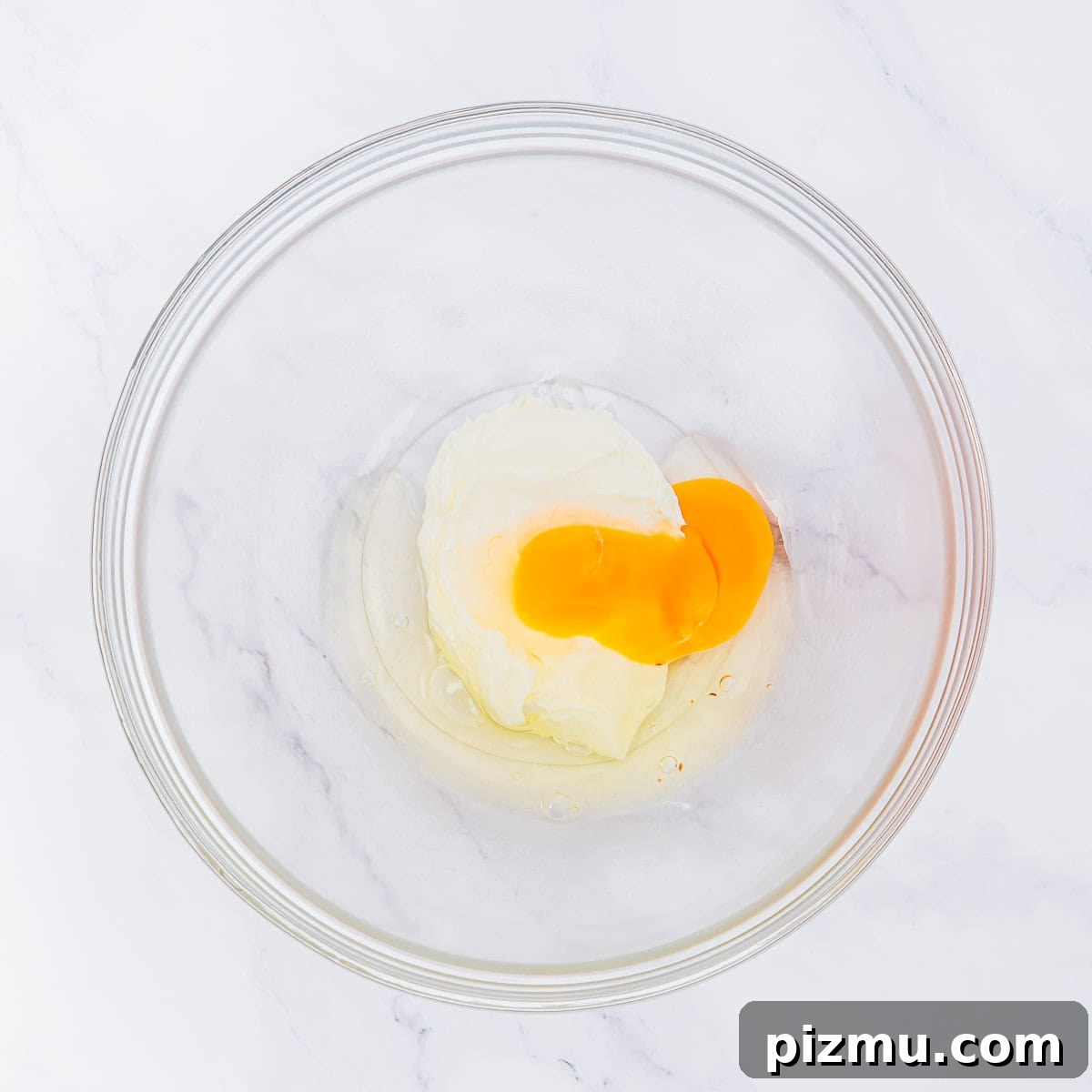 Flaky Almond Flour Biscuits Recipe 5 A glass mixing bowl containing cream cheese and raw eggs on a white marble surface.