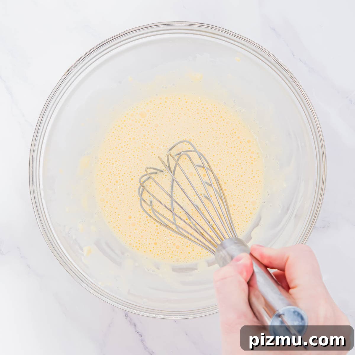 Flaky Almond Flour Biscuits Recipe 6 A hand is holding a whisk and mixing a pale yellow batter in a clear glass bowl on a white marble surface.