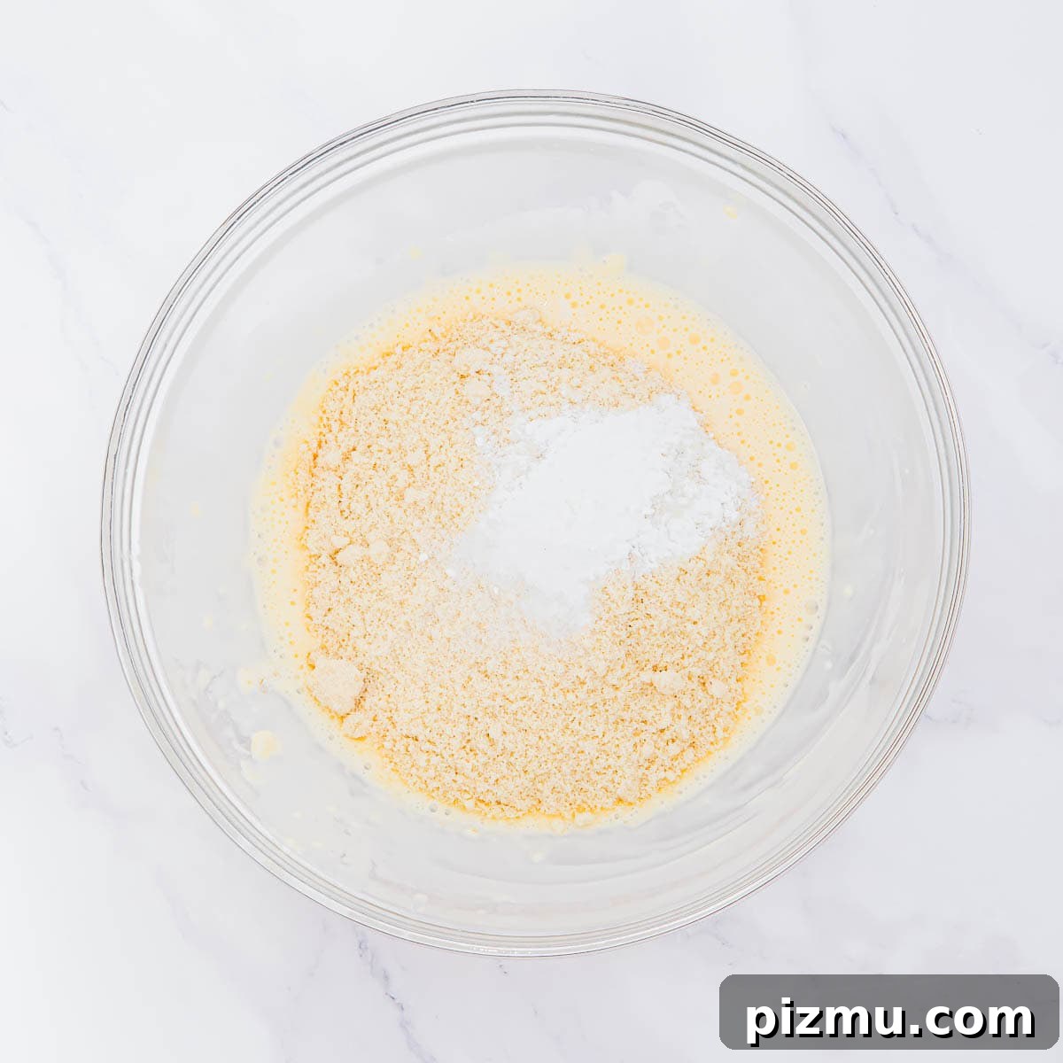 Flaky Almond Flour Biscuits Recipe 7 A clear glass bowl containing a yellow, beaten mixture with dry almond flour and a mound of white baking powder on top, set on a white marble surface.