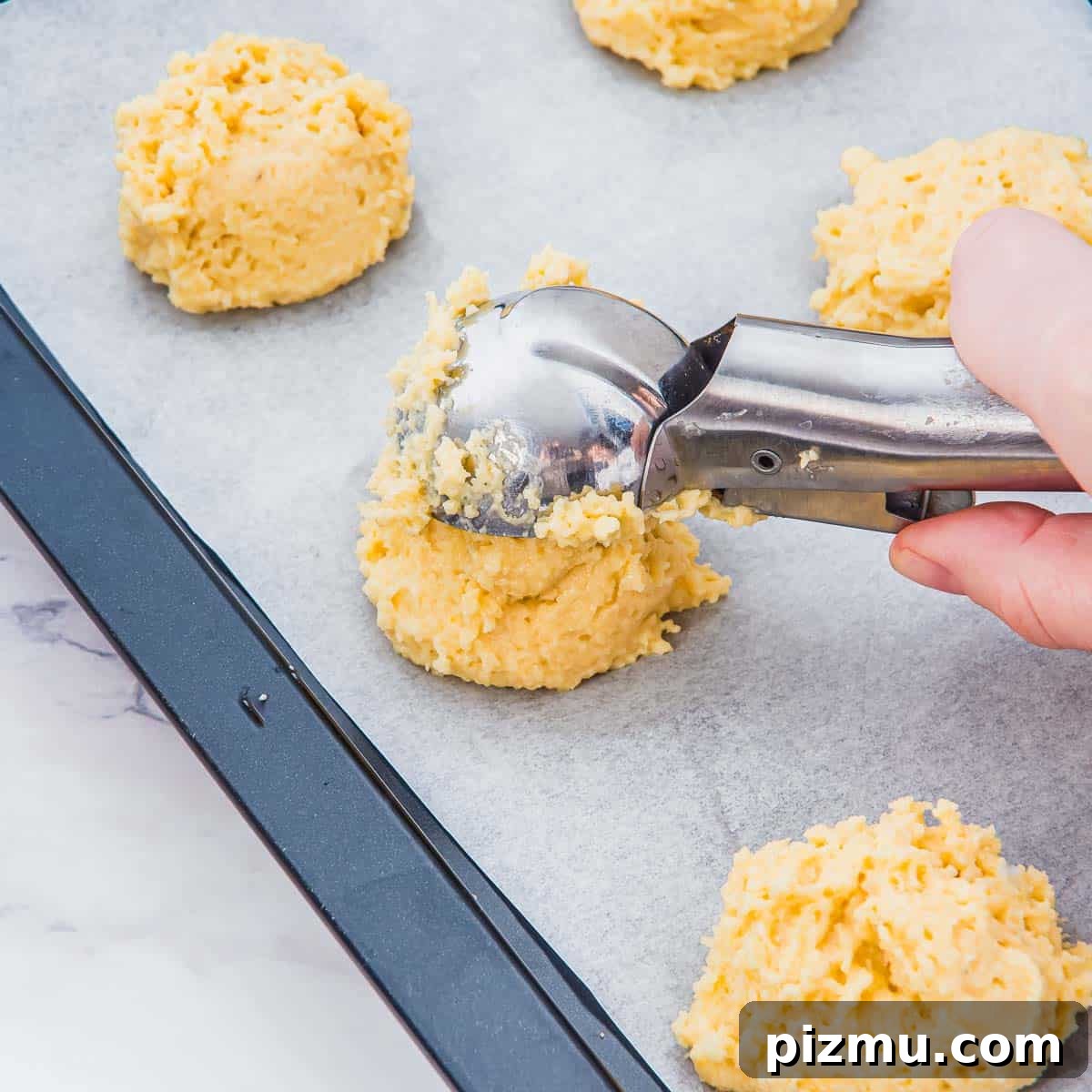Flaky Almond Flour Biscuits Recipe 9 A hand uses an ice cream scoop to place balls of biscuit dough onto a parchment-lined baking tray; several dough portions are already on the tray, ready for baking.